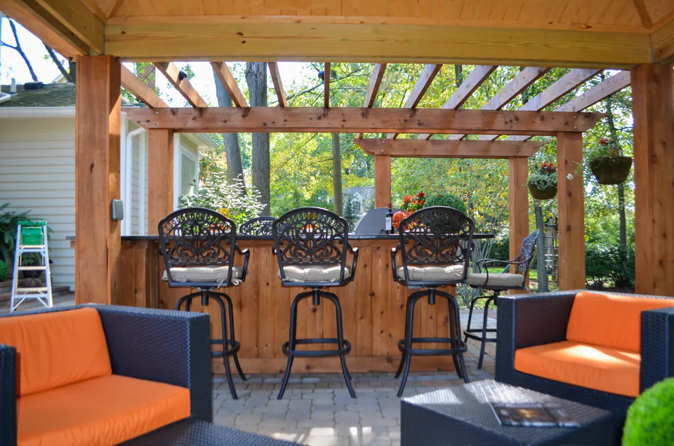 Outdoor bar seating under pergola framing with lounge seating, wood counter, paver patio, and open backyard.